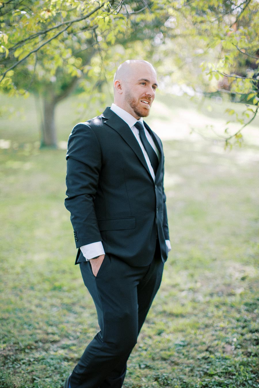 A man in a formal black suit standing outside on a sunny day, surrounded by greenery and soft focus trees in the background.