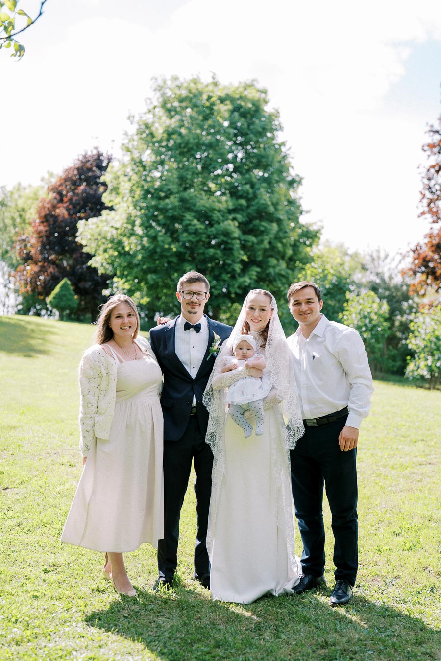 Family posing outdoors at a celebration, featuring adults in formal attire and a baby dressed in white, with a lush green park backdrop.