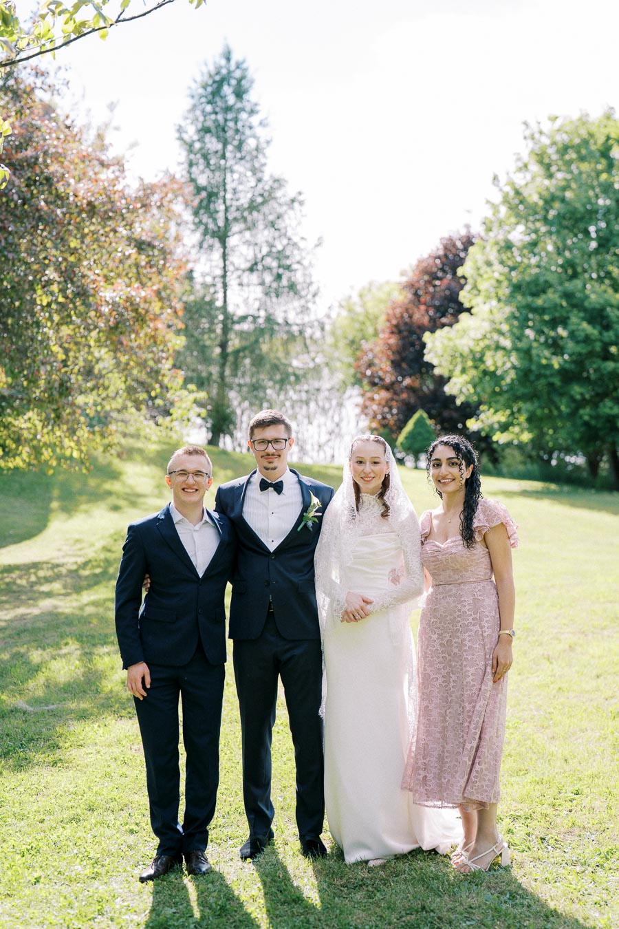 A group of four people dressed in formal attire standing together on a sunny day in a lush, green garden setting.