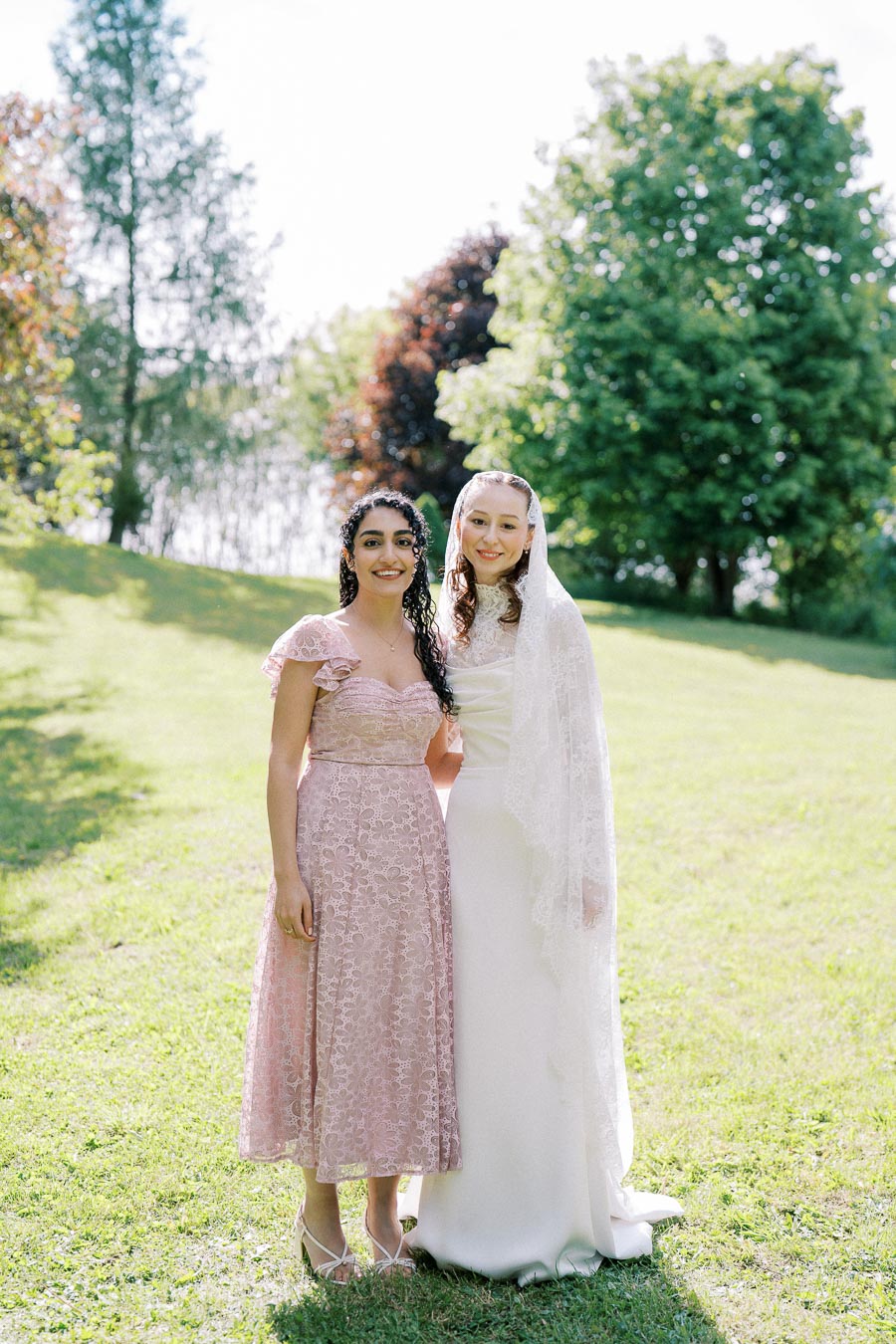 Two women standing outdoors on a sunny day, with one wearing a wedding dress and the other in a pink lace dress, smiling in a lush green park setting.