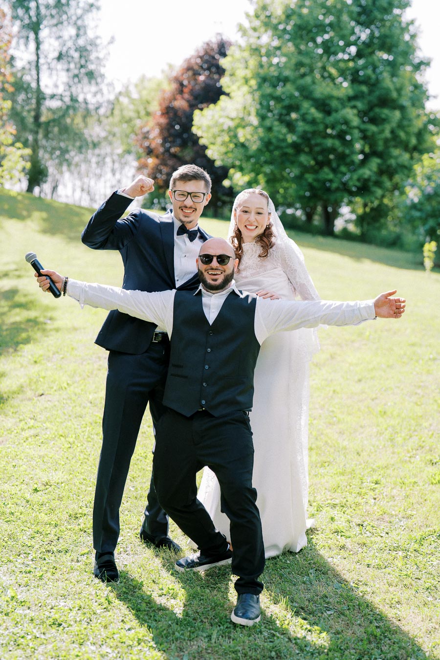 A joyful bride and groom pose with their enthusiastic groomsman holding a microphone at an outdoor wedding ceremony, surrounded by lush greenery and sunlight.