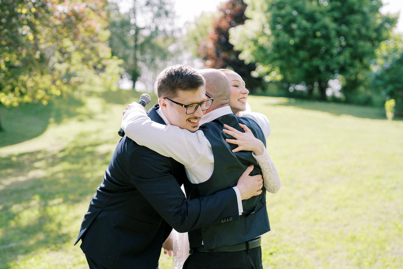 A joyful outdoor wedding ceremony with two men and a woman embracing in celebration under a bright, sunny sky surrounded by lush greenery.