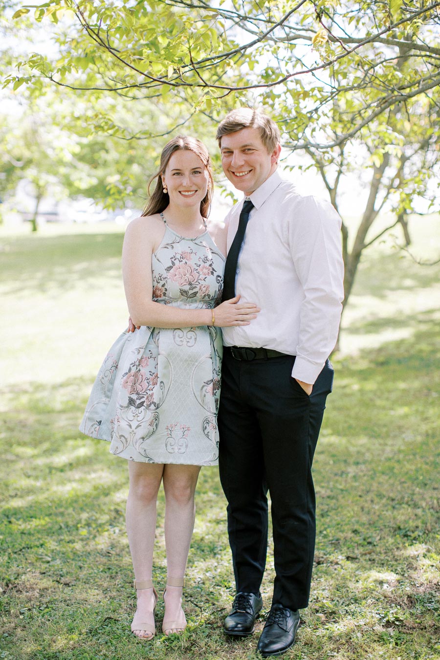 A smiling couple standing together in a sunlit park, with green trees and grass in the background. The woman is wearing a floral dress, while the man is dressed in a white shirt and black pants.