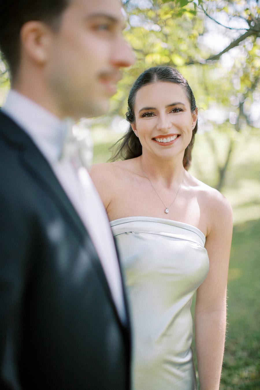 A woman smiling in a white strapless dress standing outdoors with a blurred person in a suit in the foreground. The scene is set in a lush, green natural environment with sunlight filtering through the trees.