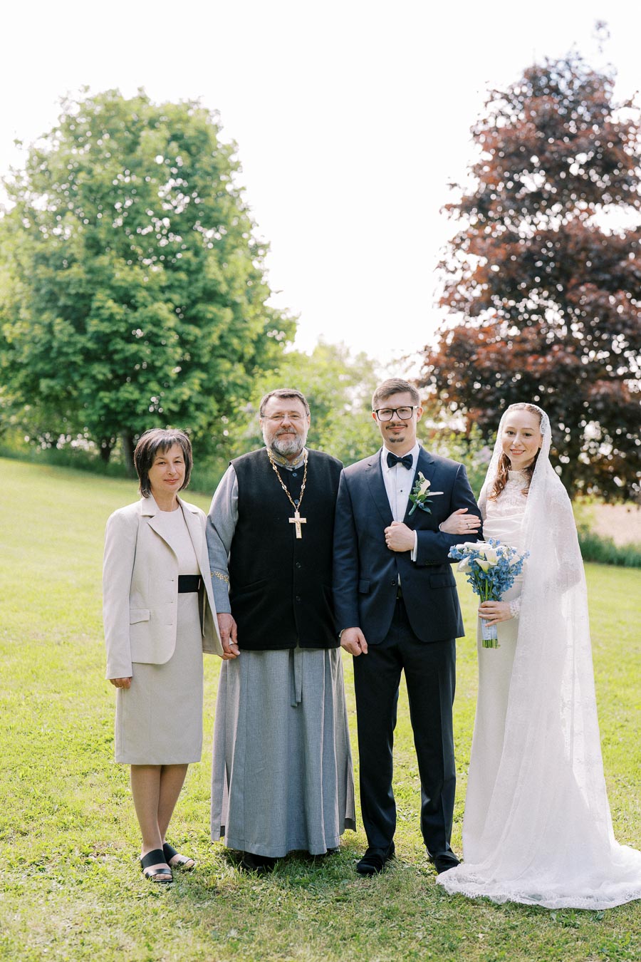 A wedding group photo with a bride in a white dress and veil holding a bouquet, a groom in a suit with a boutonniere, a religious officiant with a clerical robe and cross, and a woman in a beige outfit, set outdoors in a green park.