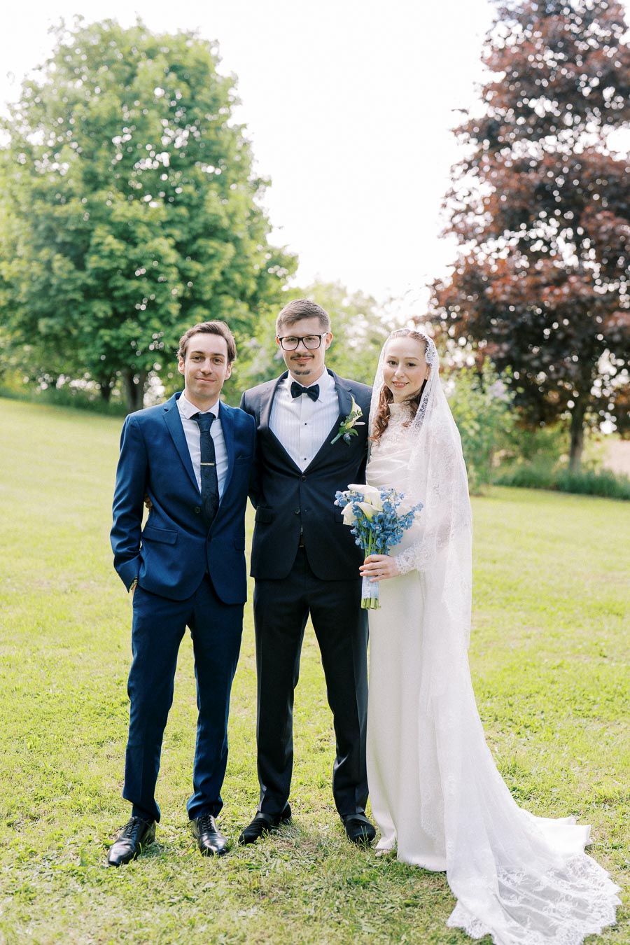 Elegant wedding portrait featuring a bride in a lace gown holding a bouquet, standing beside two groomsmen in tailored suits, on a sunny day in a lush green park setting.