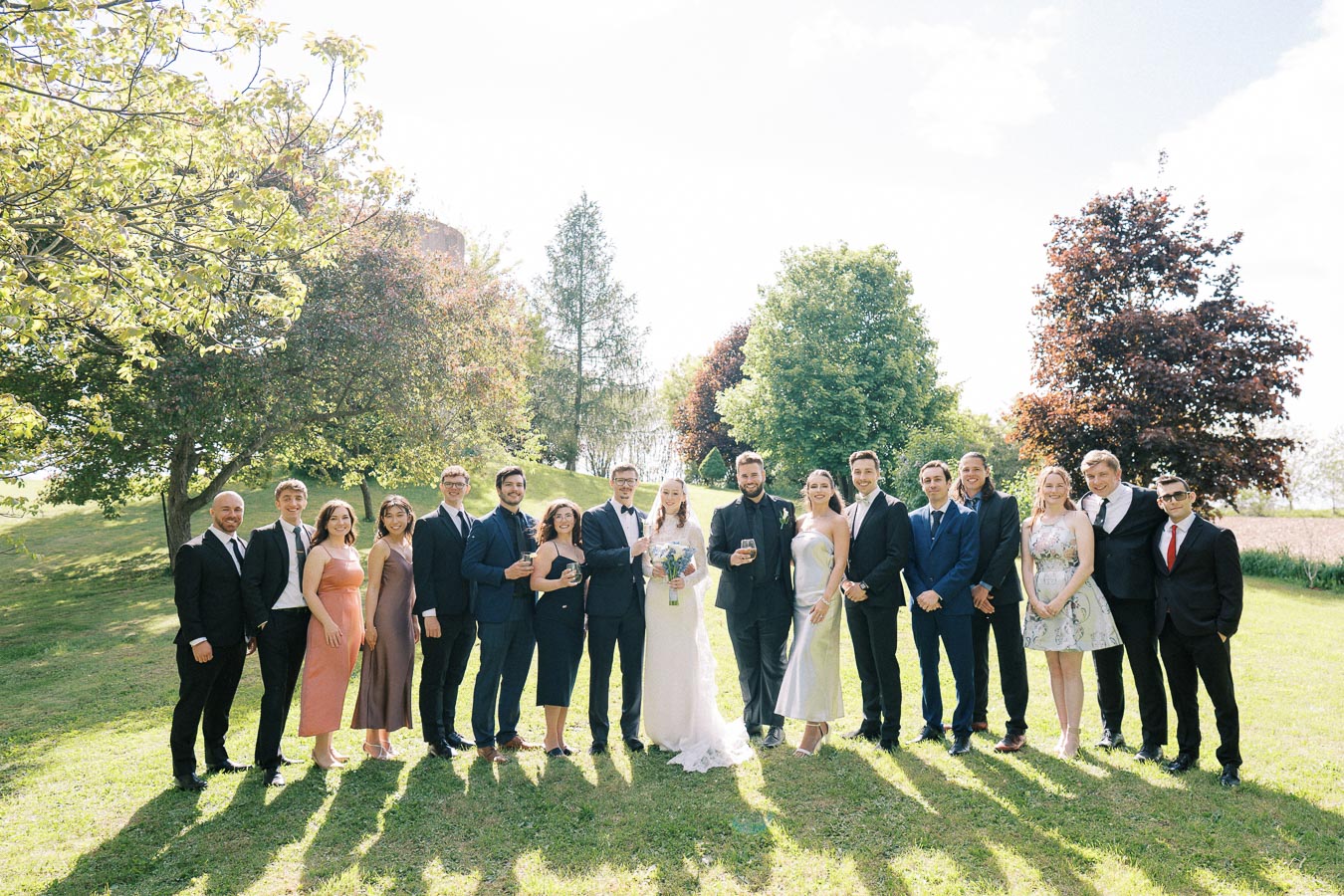 A diverse group of elegantly dressed people standing together outdoors in a sunny park, celebrating a wedding. The group includes the bride in a white gown and the groom in a suit, surrounded by friends and family, with green trees and blue sky in the background.