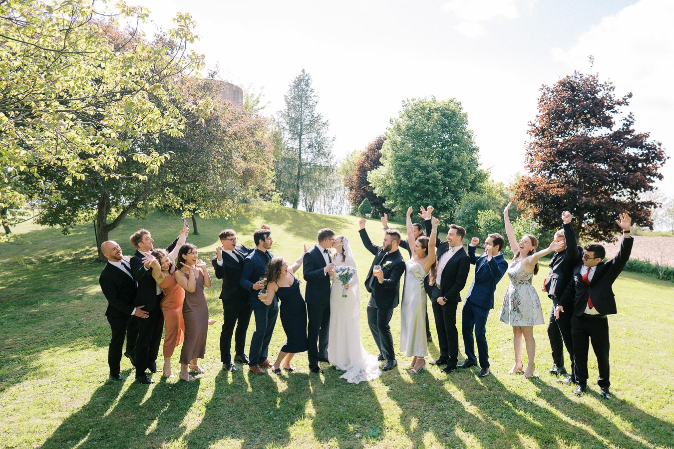 A joyful wedding party celebrating outdoors, featuring a bride and groom surrounded by friends and family, all dressed in formal attire and raising their hands in excitement, set against a lush green park backdrop with trees and a bright blue sky.