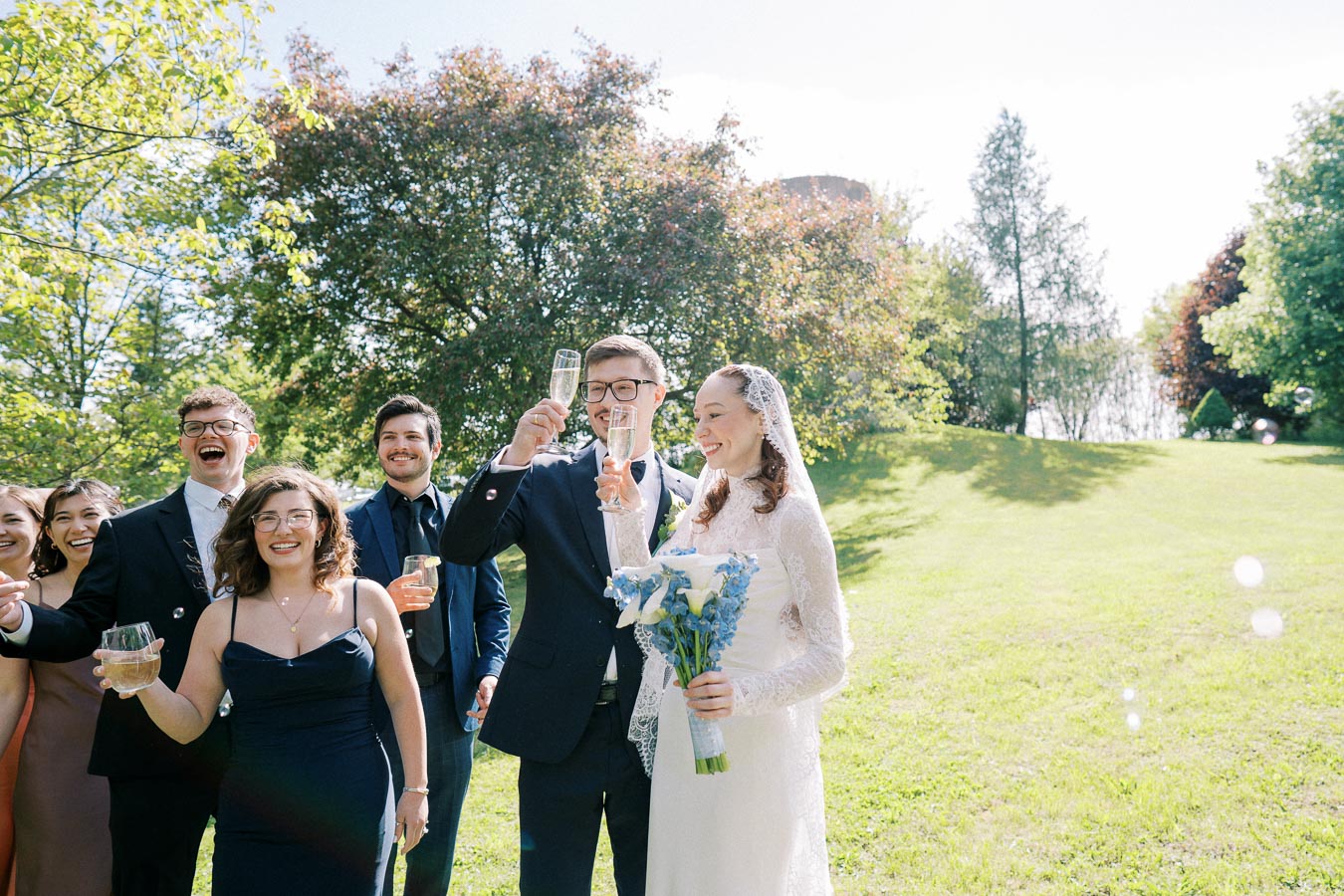 A joyful wedding party celebrating outdoors, featuring a bride in a lace gown holding a blue and white bouquet and a groom in a suit toasting with guests in formal attire, with lush green trees and sunlight in the background.