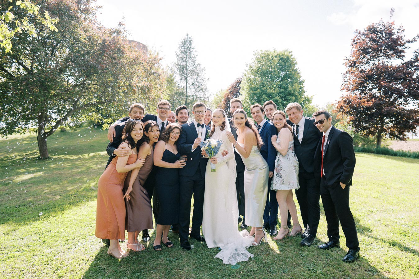 A joyful wedding party poses outdoors, featuring the bride in a white dress holding a bouquet, surrounded by bridesmaids in elegant dresses and groomsmen in suits on a sunny day with lush green trees in the background.