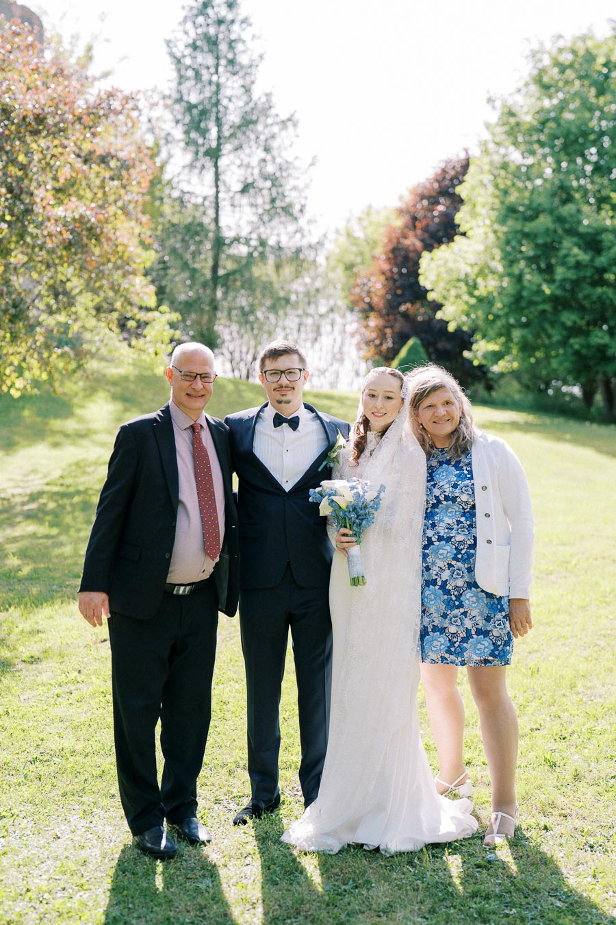 A bride and groom pose with two family members outdoors on a sunny day, surrounded by lush green trees and grass, during a wedding celebration.