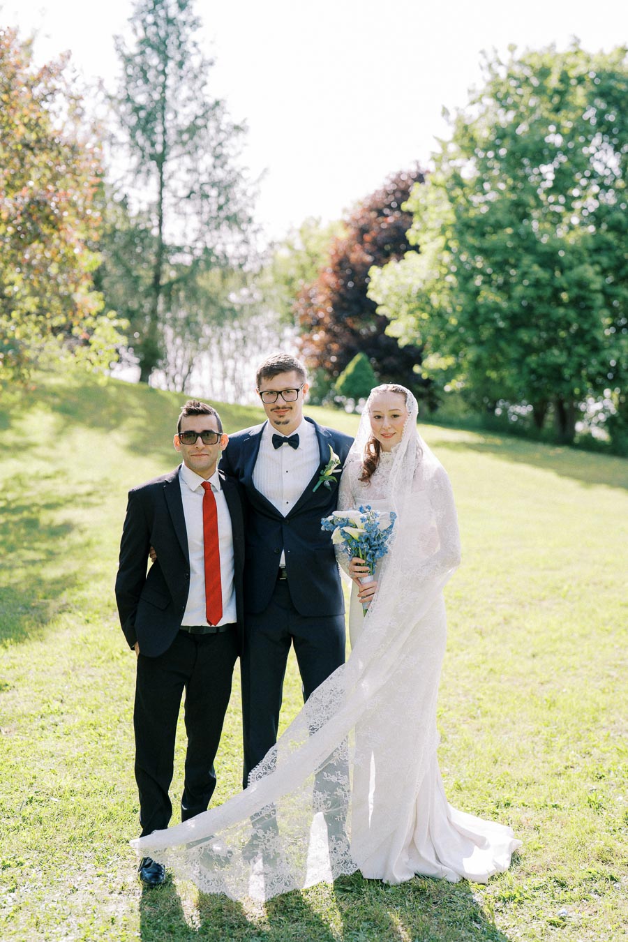 A bride in a white lace gown holding a bouquet stands beside two men in suits on a sunlit, grassy field with trees in the background.