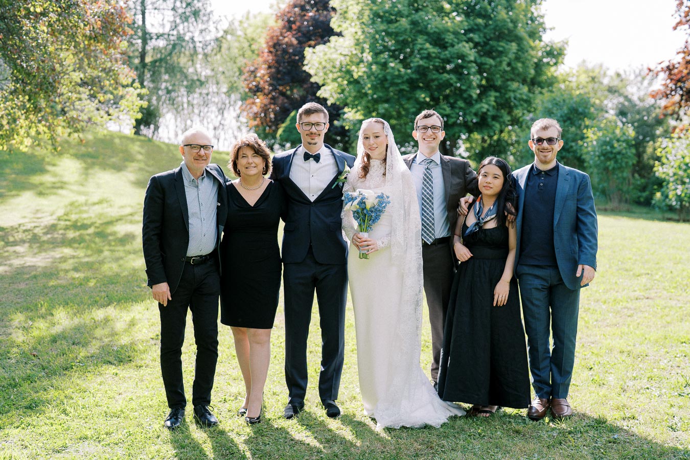 Wedding group photo in a sunlit garden, featuring a bride in a white gown holding a bouquet, standing beside a groom in a suit, surrounded by family members dressed in formal attire amidst lush greenery.