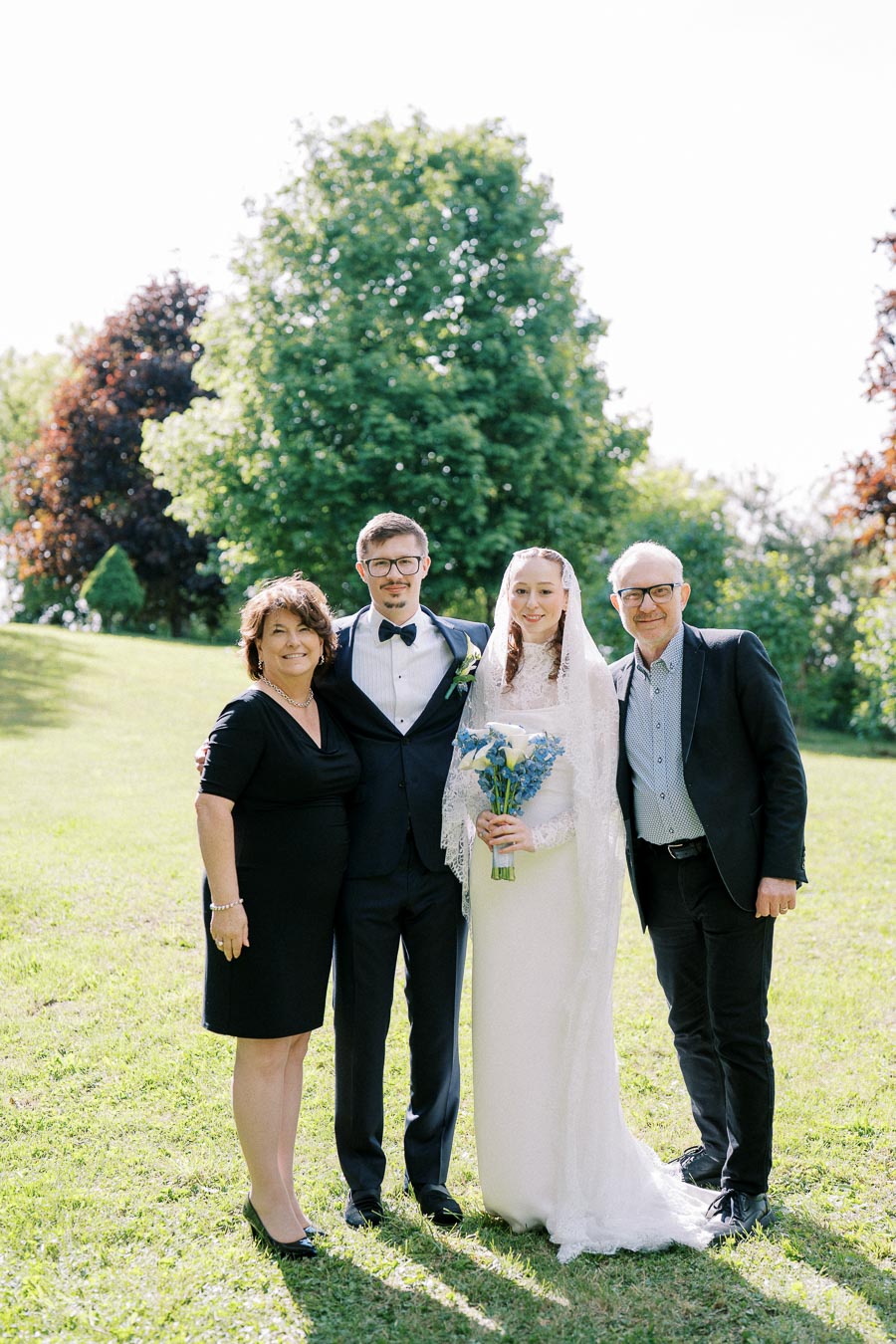 A wedding couple and two guests standing on a grass lawn with trees in the background, the bride holding a bouquet of blue flowers.