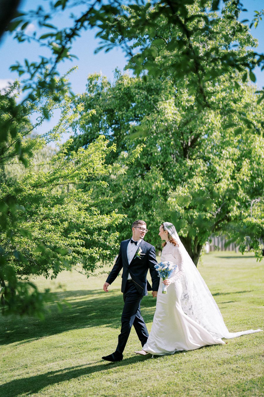 A bride and groom walk hand in hand through a lush green garden on a sunny day, with the bride in a white gown and the groom in a black tuxedo, beneath a clear blue sky.