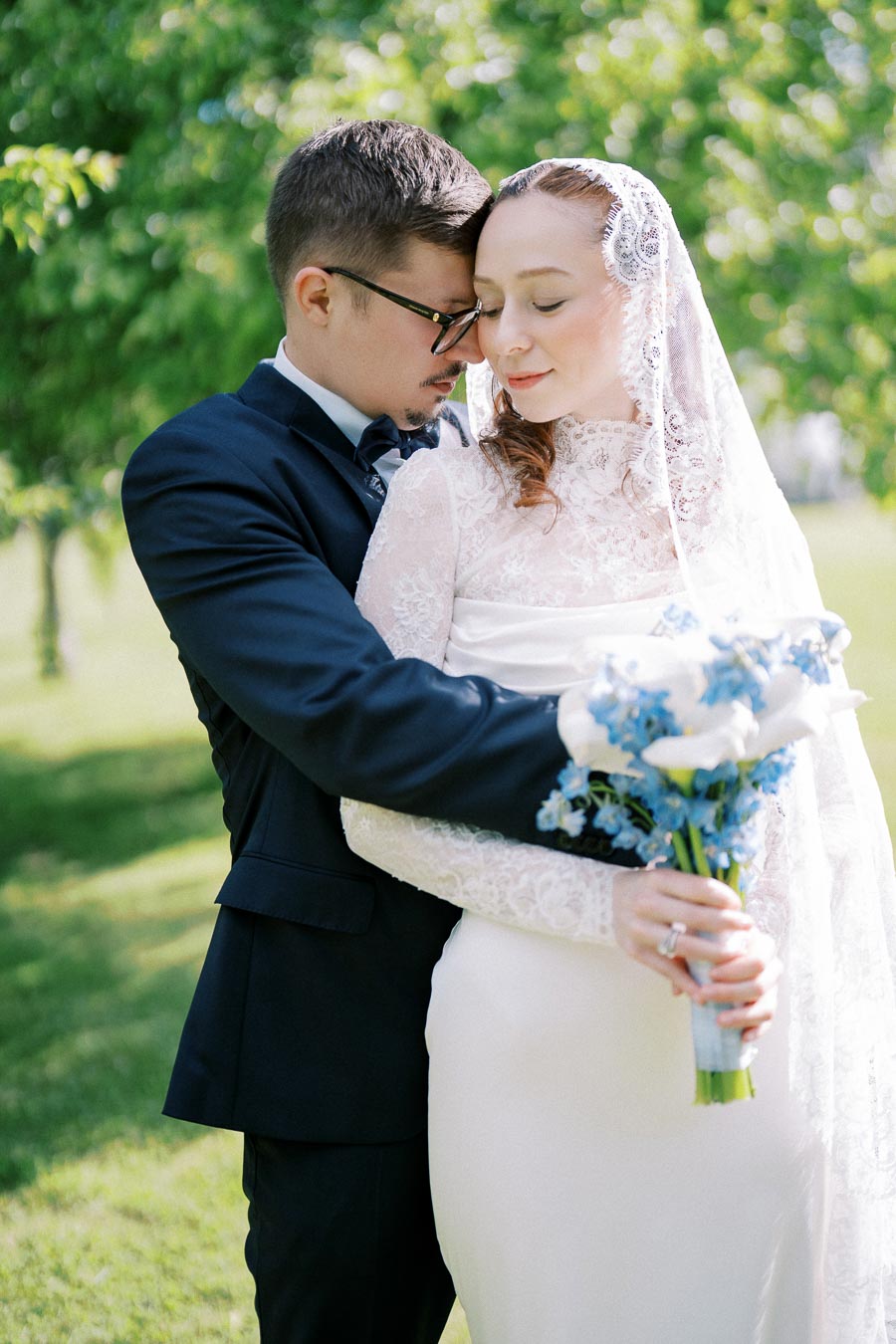 A bride and groom embrace lovingly in a sunlit garden, the bride holding a bouquet of blue and white flowers, wearing a lace dress and veil, while the groom wears a dark suit and glasses.