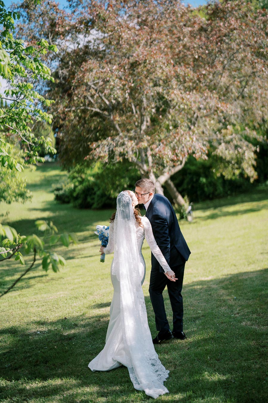 A newlywed couple sharing a kiss outdoors, with the bride in a long white dress and veil holding a bouquet of blue flowers, set against a lush green landscape with trees.