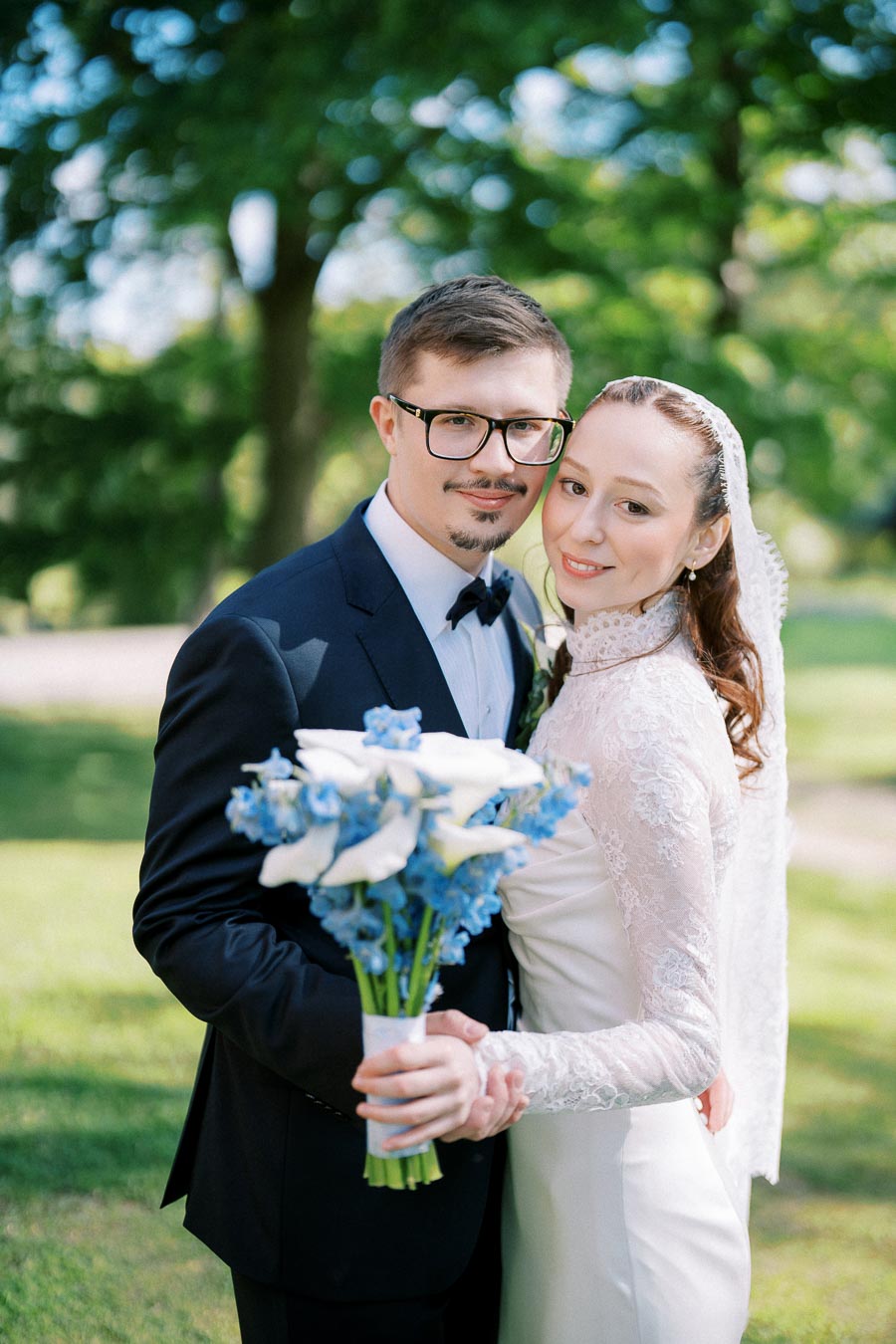A newlywed couple poses for a wedding portrait in a lush green park, with the groom in a black suit holding a bouquet of white and blue flowers, and the bride in a white lace gown smiling warmly.
