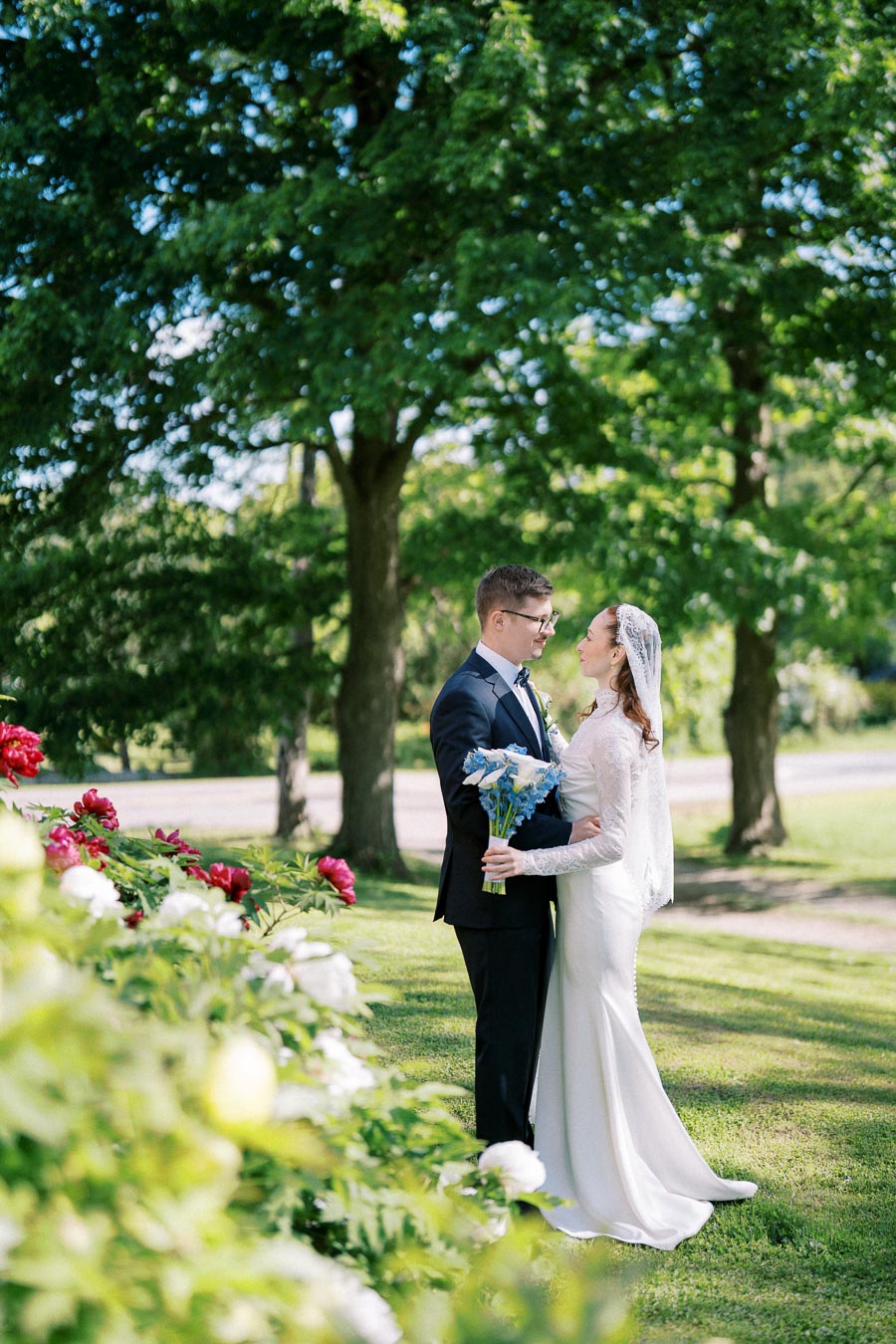 A bride and groom embracing in a garden setting, surrounded by lush green trees and vibrant flowers, with the bride holding a bouquet of blue flowers.