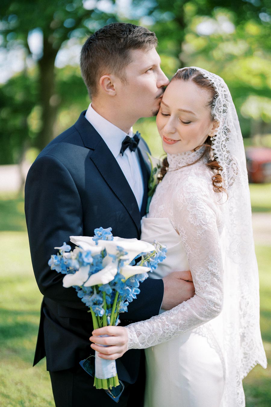 Wedded couple embracing outdoors, groom in navy suit and bride in lace gown holding blue and white bouquet, surrounded by lush green foliage.