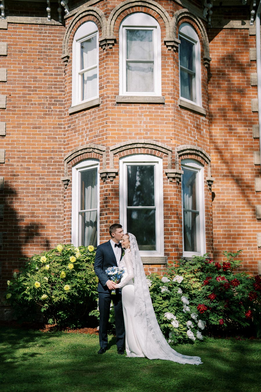A bride and groom stand in an embrace in front of a vintage brick building, surrounded by vibrant flowers and greenery. The bride wears a long white gown with a lace veil, while the groom is dressed in a dark suit and bow tie.