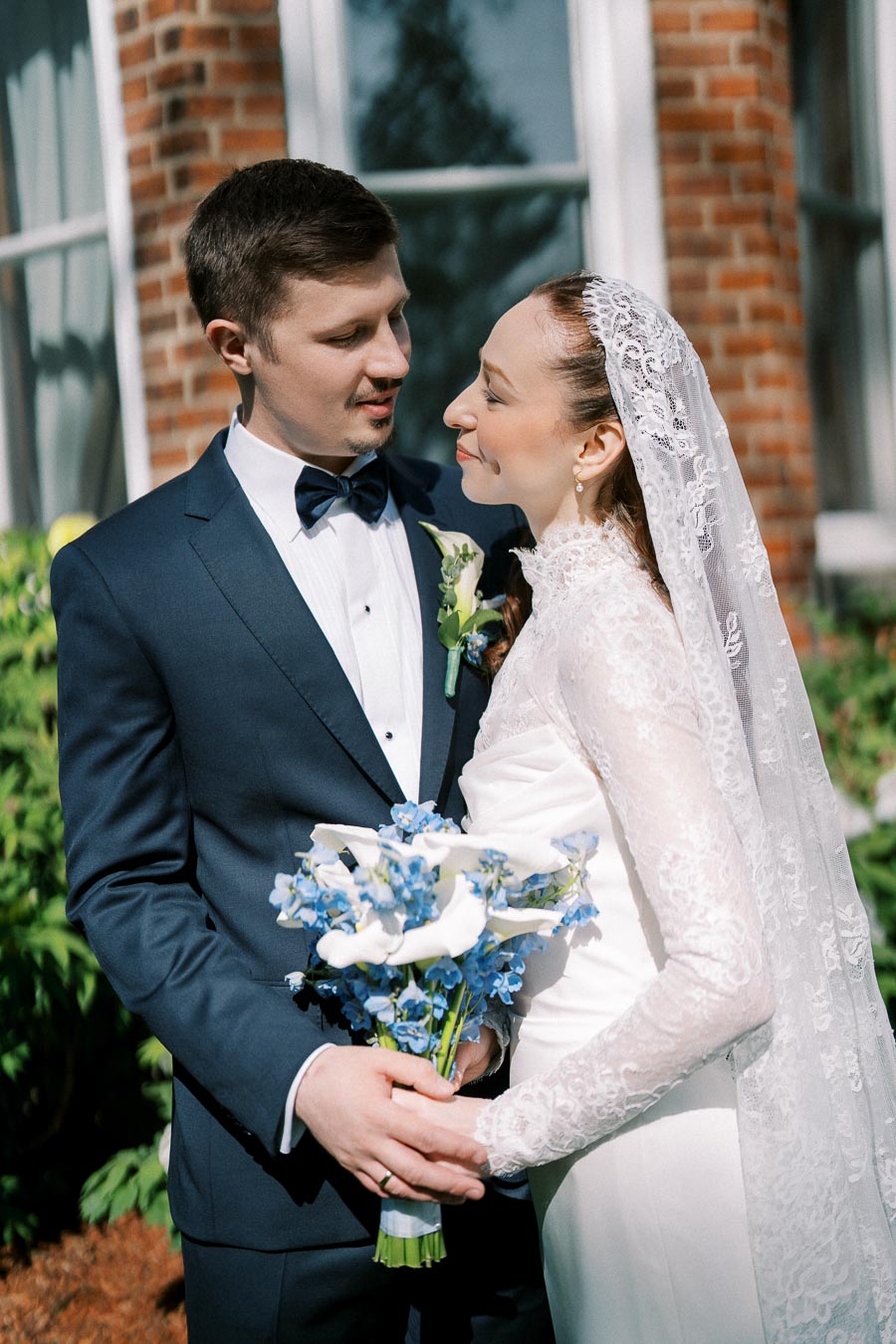 A bride and groom share an intimate moment outside a brick building, with the bride holding a bouquet of blue and white flowers. She wears a lace veil and gown, and he is dressed in a navy suit with a bow tie.