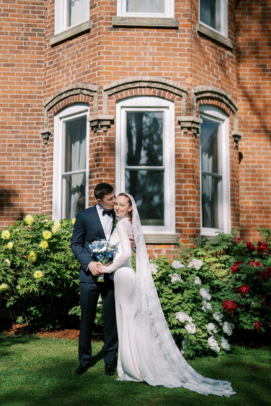 A bride in a long white dress and veil holds a bouquet of blue flowers, standing with the groom in a black suit and bow tie in front of a brick building with lush green and flowering bushes, under a clear sky.