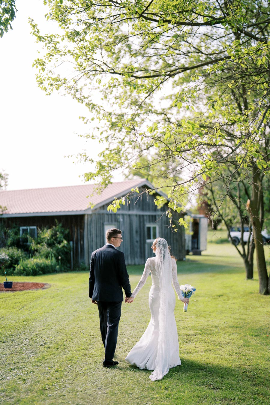 A bride and groom holding hands walk towards a rustic barn in a lush, green outdoor setting. The bride is in an elegant white gown with a long veil, and the groom is in a classic black suit. Sunlight filters through the trees, creating a romantic and serene atmosphere.