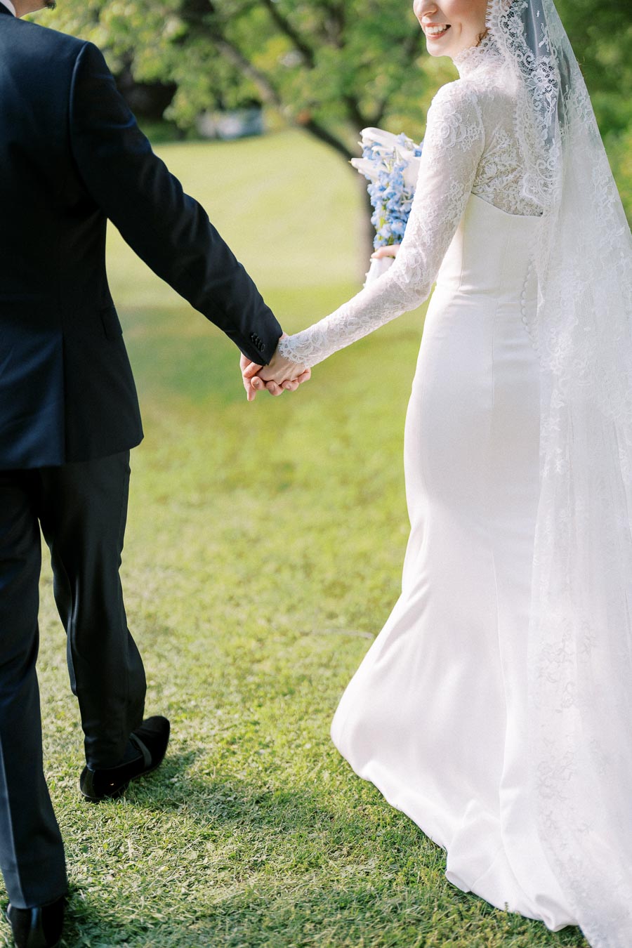 Bride and groom holding hands, walking on a grassy field, with the bride wearing a lace wedding dress and veil while holding a bouquet.