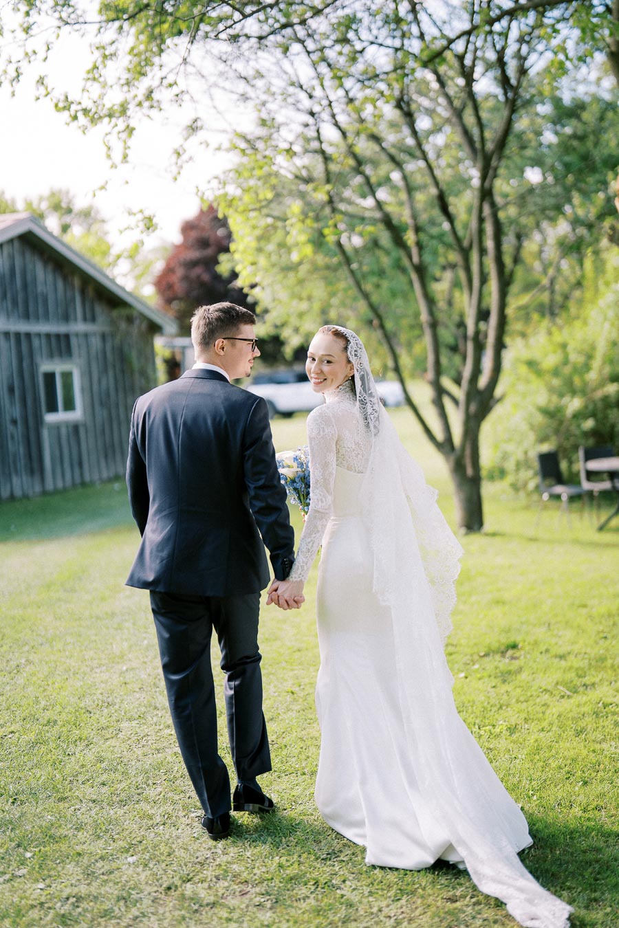Bride and groom walking hand in hand on a sunny day, surrounded by lush greenery and a rustic barn, showcasing a picturesque outdoor wedding scene.