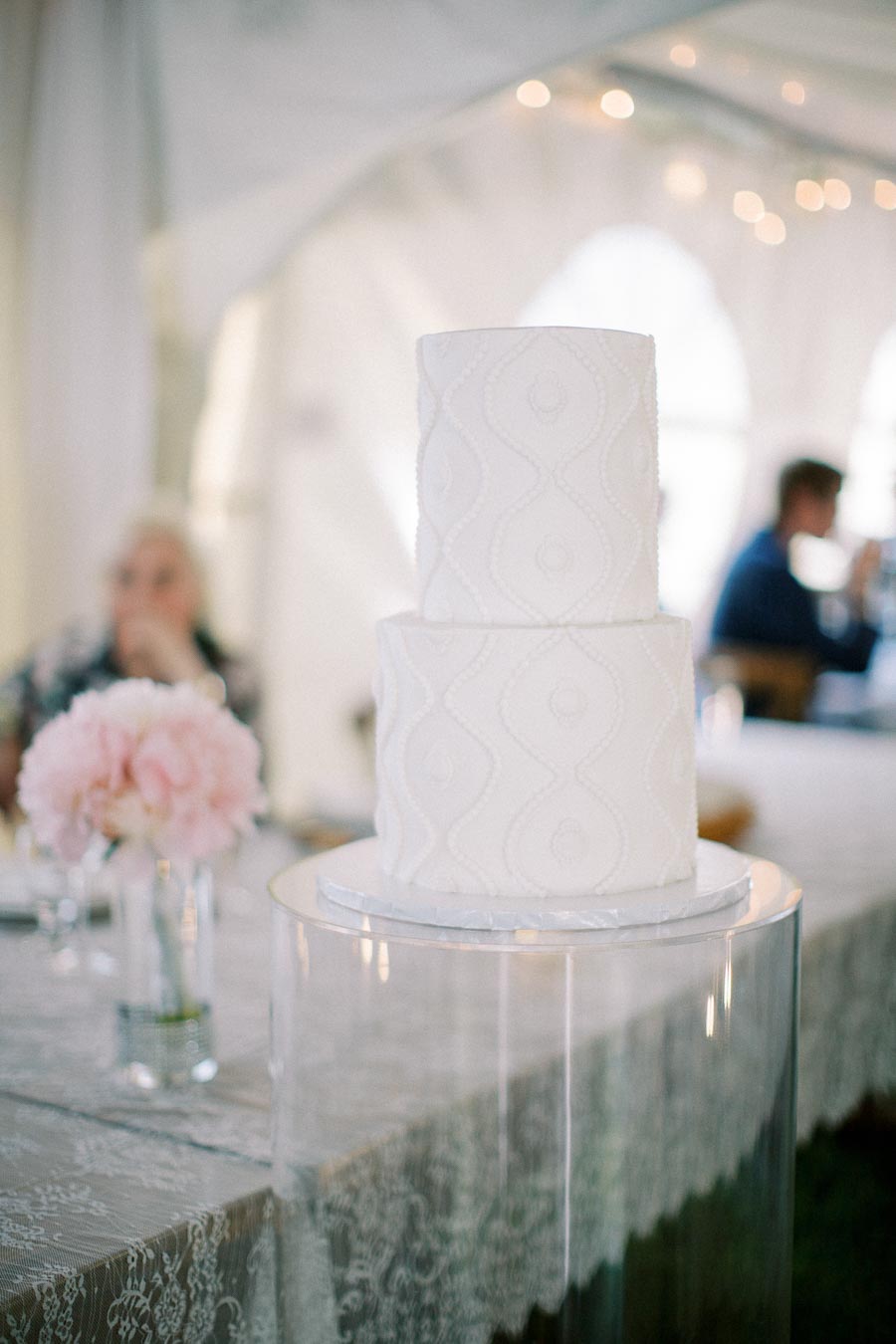 Elegant two-tier white wedding cake with intricate patterns on glass stand, set in a beautifully decorated reception with soft floral arrangements and lace tablecloth.