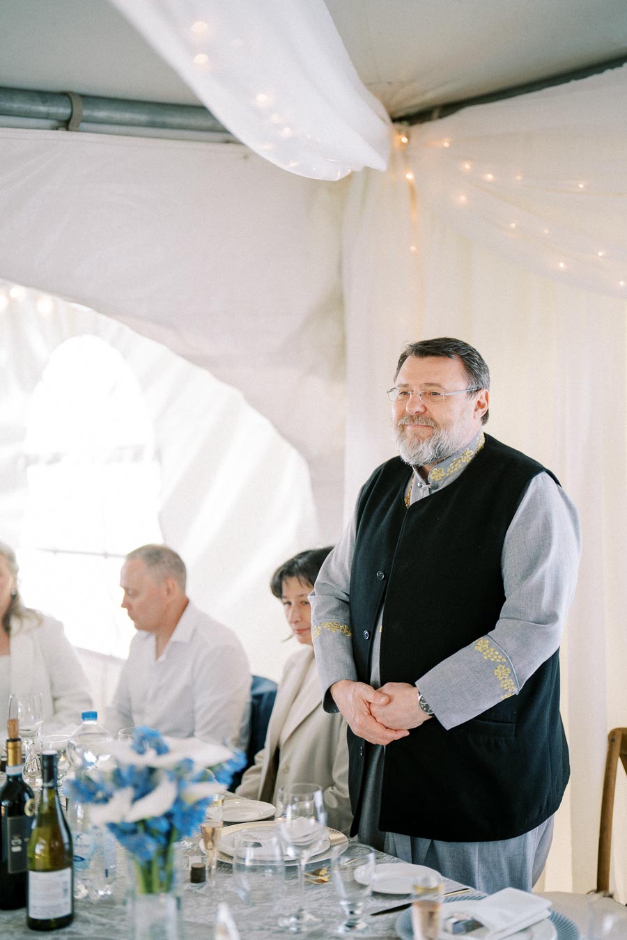 A man standing and giving a speech at a wedding reception, dressed in a traditional outfit with decorative embroidery. Guests seated at a table with blue floral centerpiece and wine bottles in a softly lit tent venue.