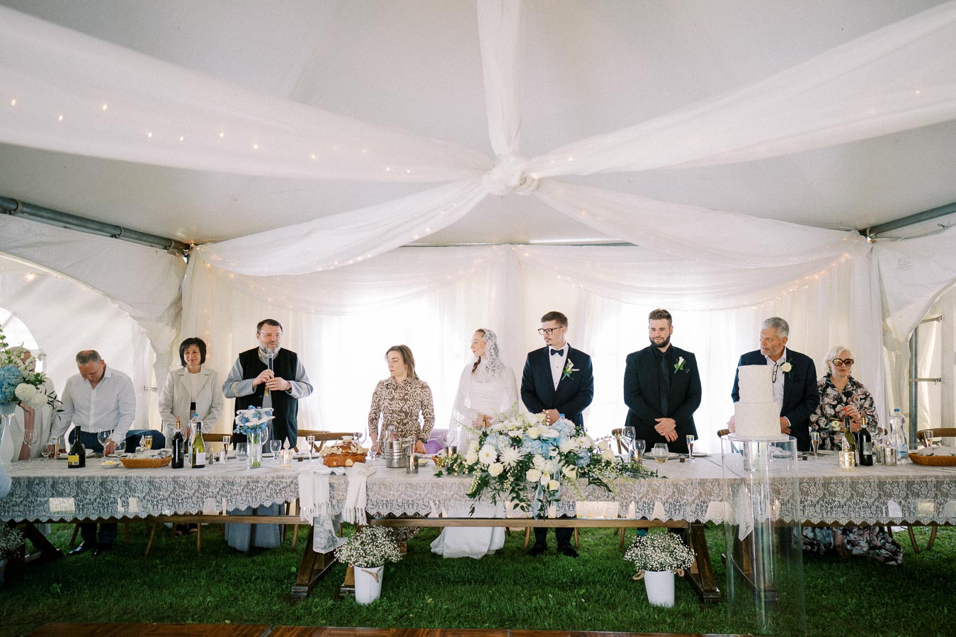 Wedding reception under a decorated tent with a long table adorned with floral arrangements. A bride, groom, and guests stand together, creating a celebratory and elegant atmosphere.