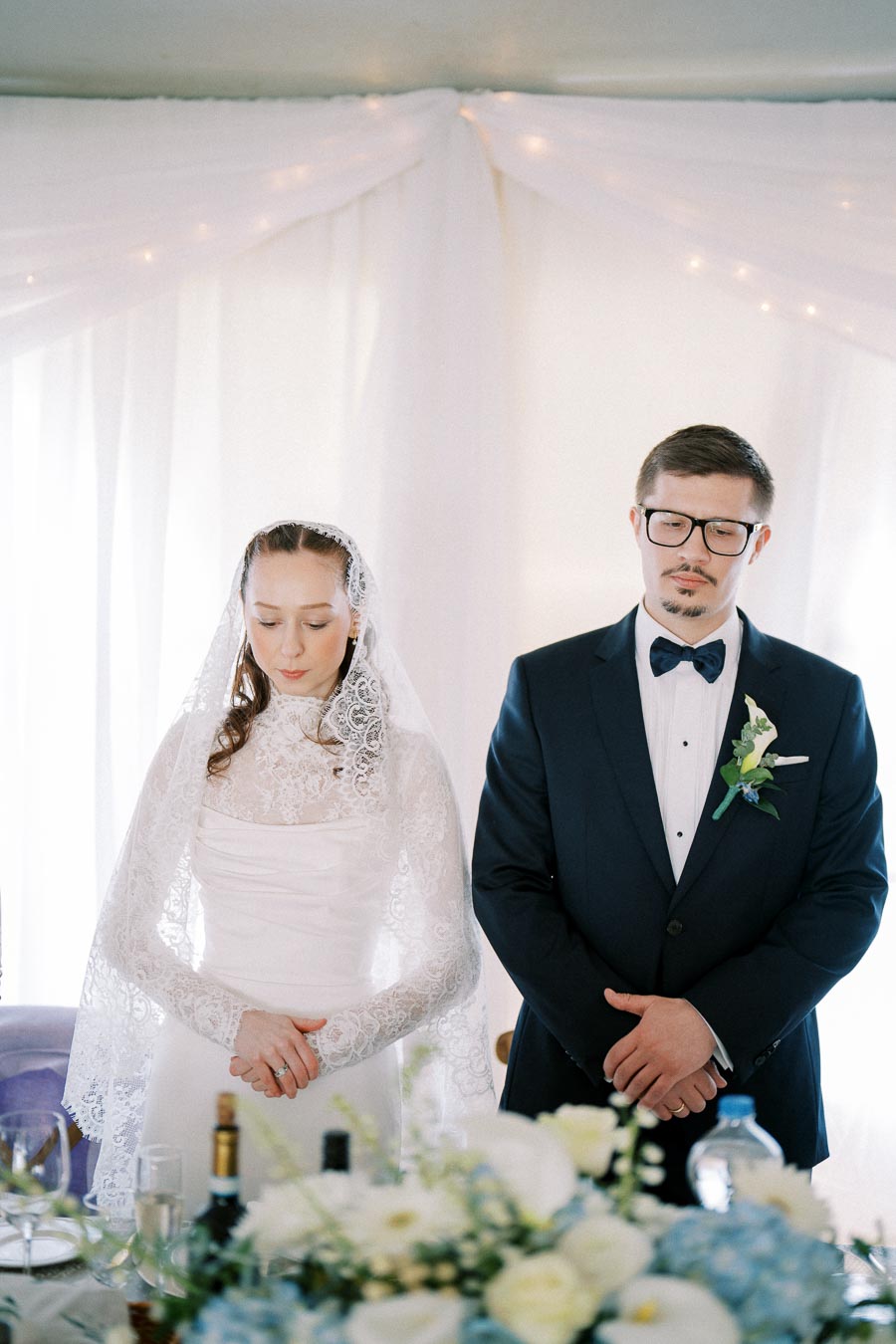 Elegant bride and groom standing together at wedding reception, adorned in white lace dress and navy blue suit with boutonniere, in a softly lit venue decorated with white drapery and floral arrangements.