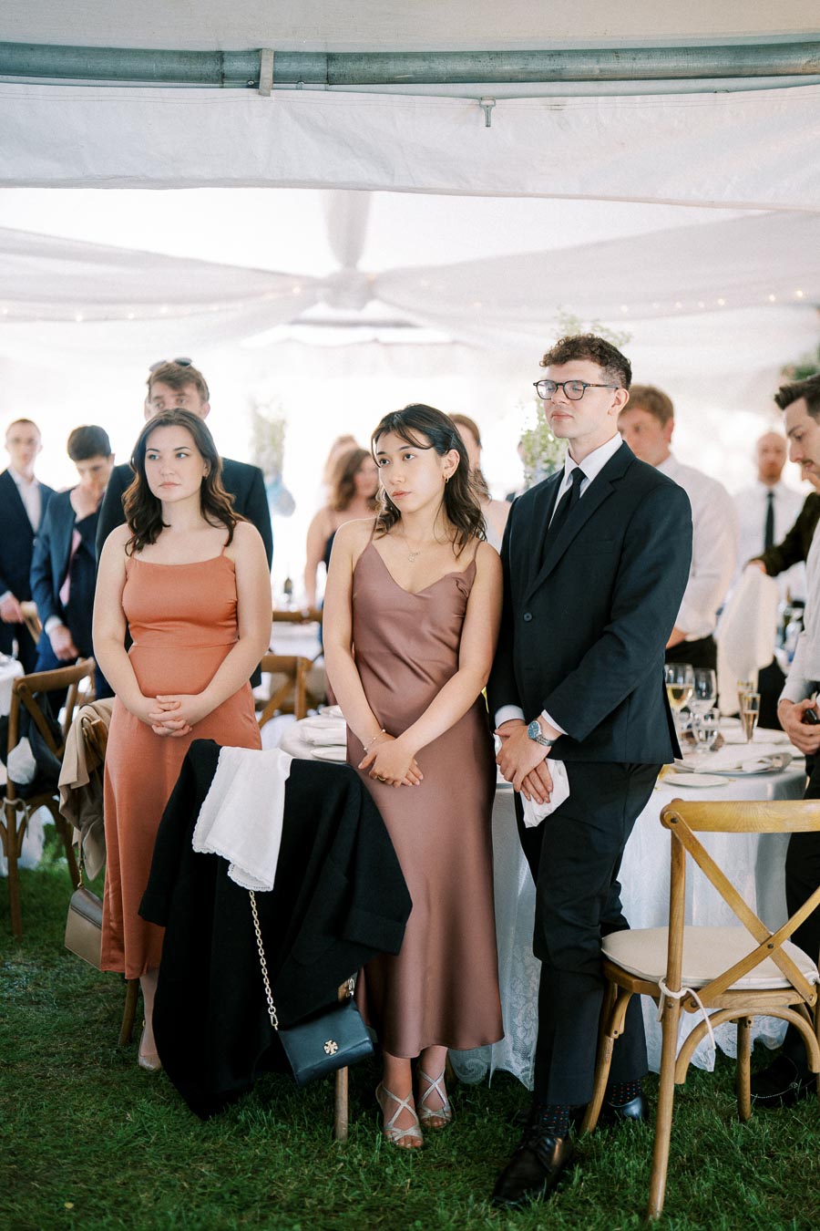 A group of elegantly dressed guests standing at a formal event, with a white canopy overhead and tables set with fine dinnerware in the background.