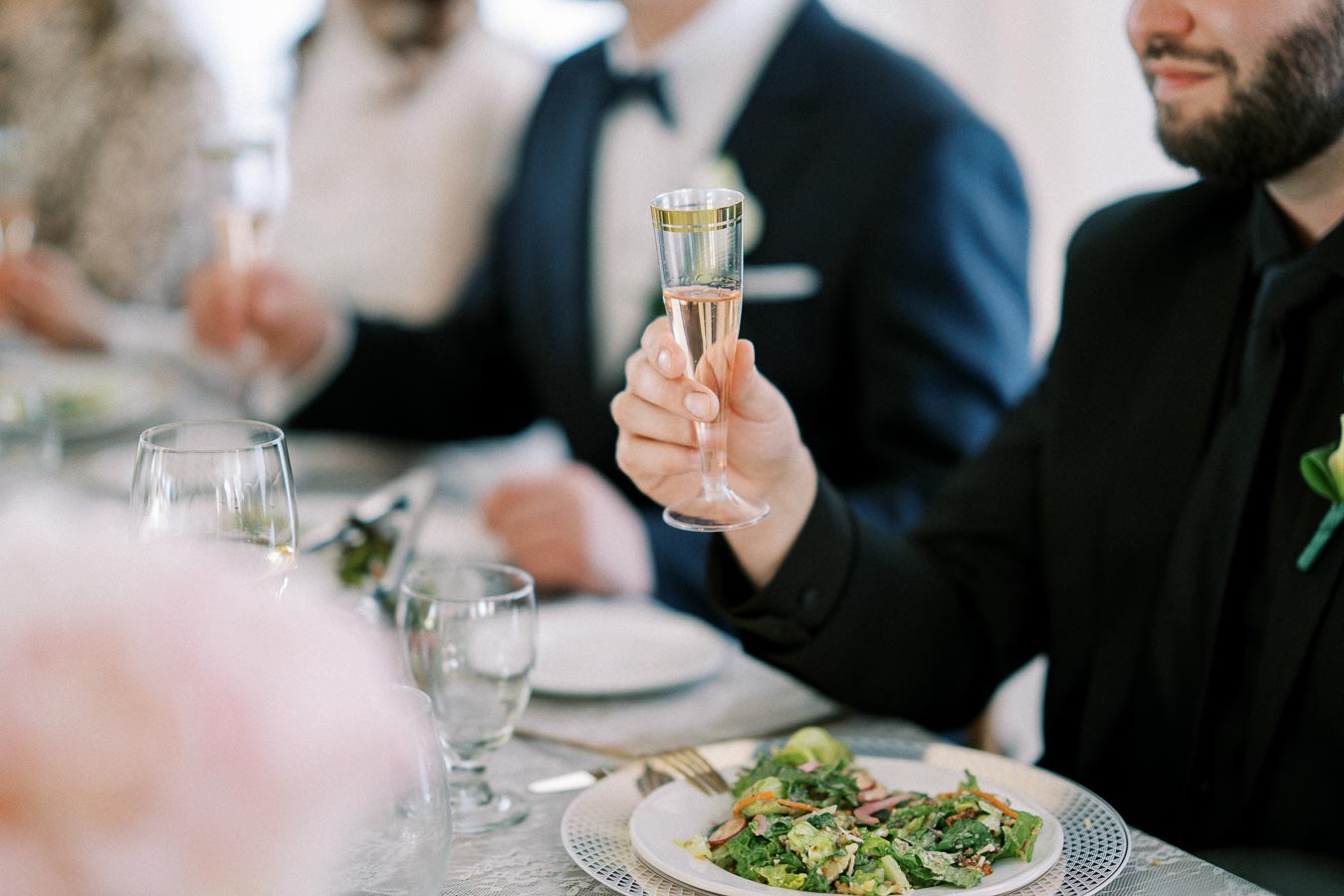 A person in formal attire holding a glass of champagne at a wedding reception table set with salad and elegant tableware.