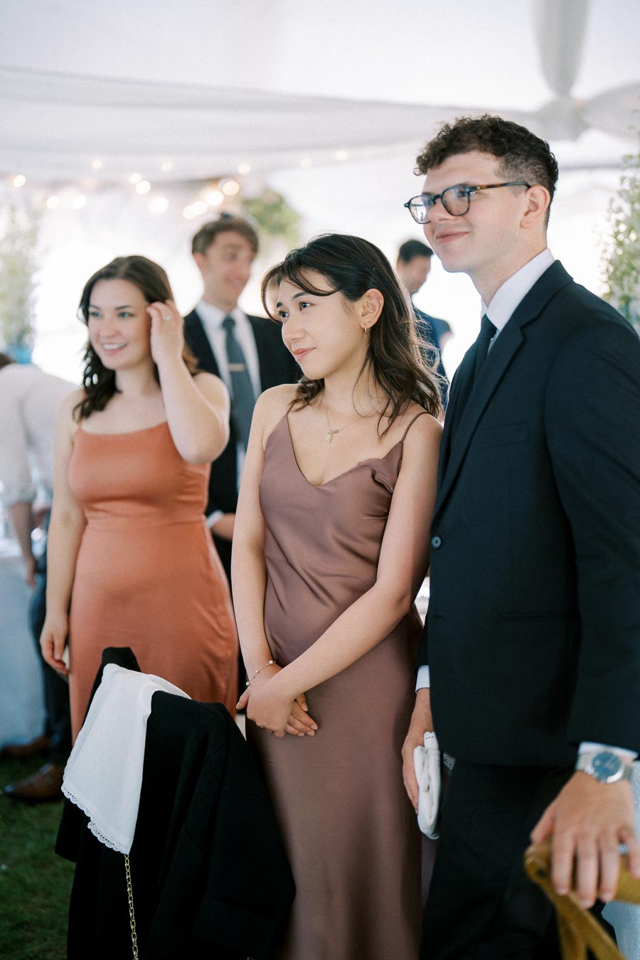 Group of elegantly dressed people attending an outdoor event, women in satin dresses and men in suits, under a decorated canopy with soft lighting.