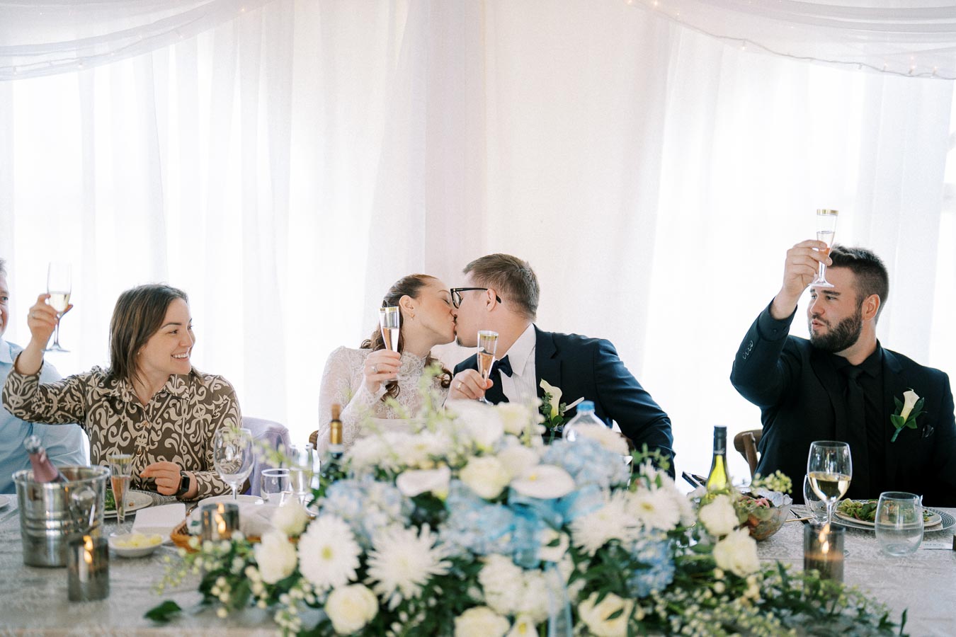 Wedding celebration with guests toasting champagne, bride and groom kissing at head table adorned with white and blue flowers, joyful atmosphere.