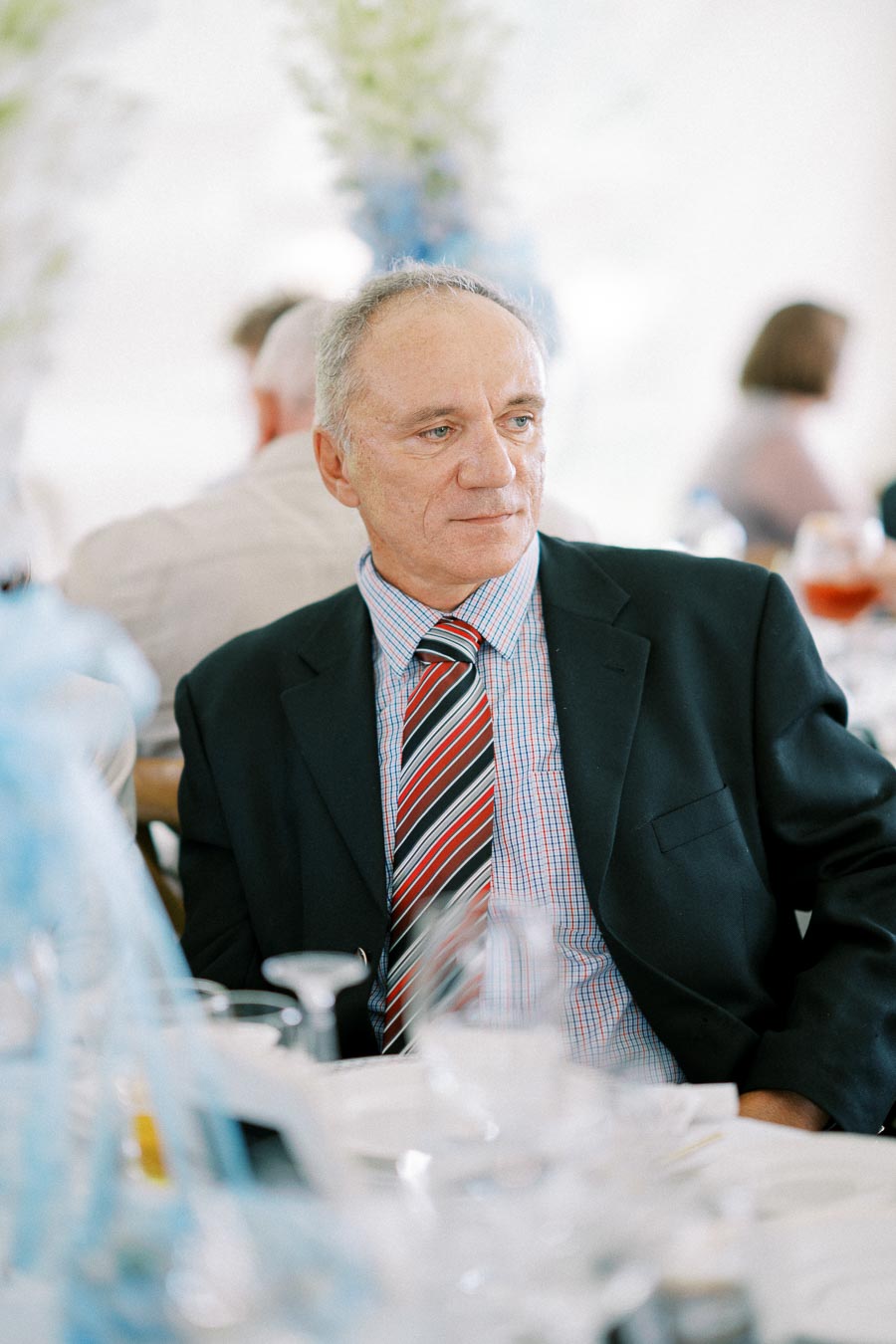 Elderly man in suit and striped tie sitting at a formal event with blurred guests in the background.
