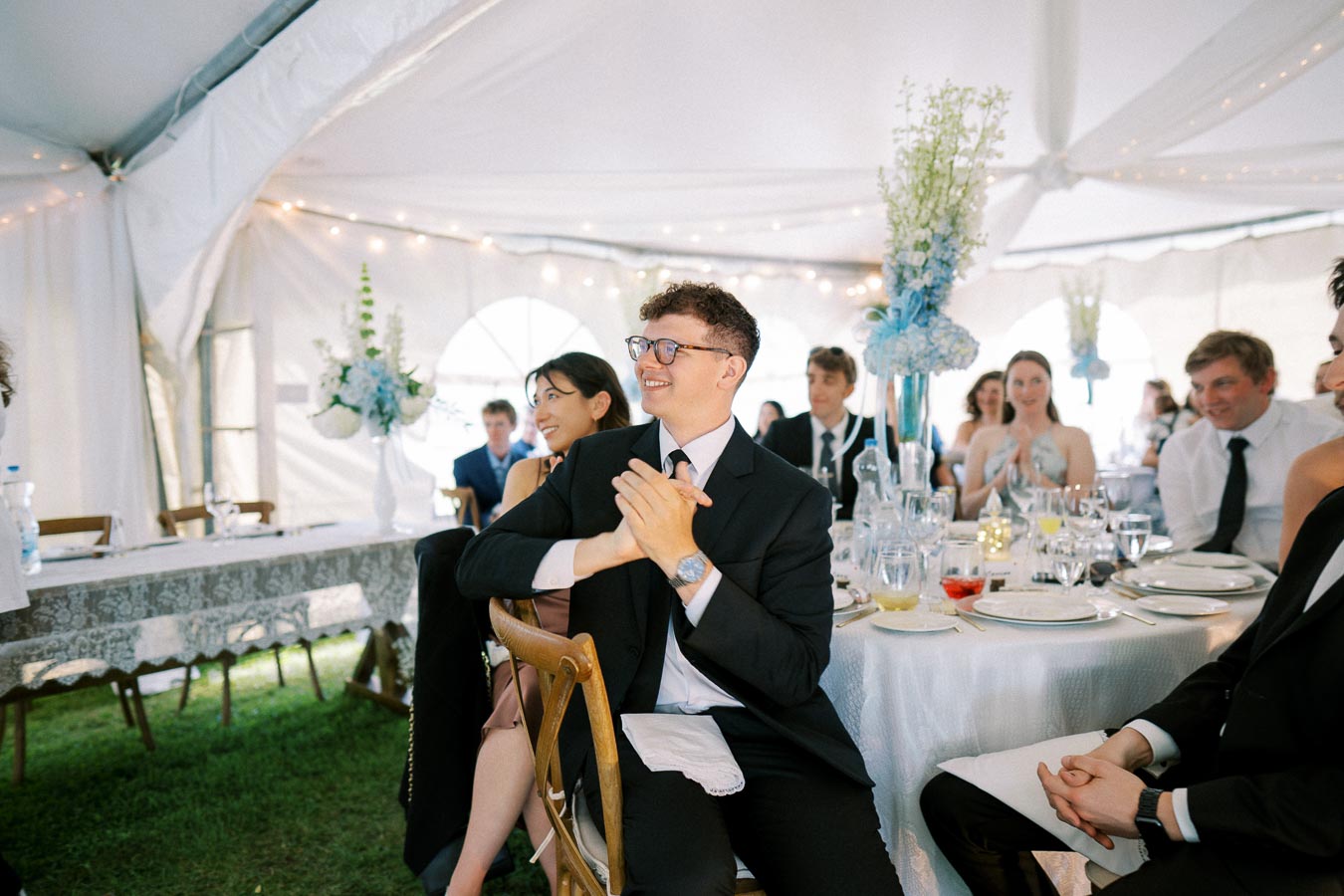 A group of elegantly dressed guests clapping and smiling at a wedding reception in a beautifully decorated white tent with floral centerpieces.