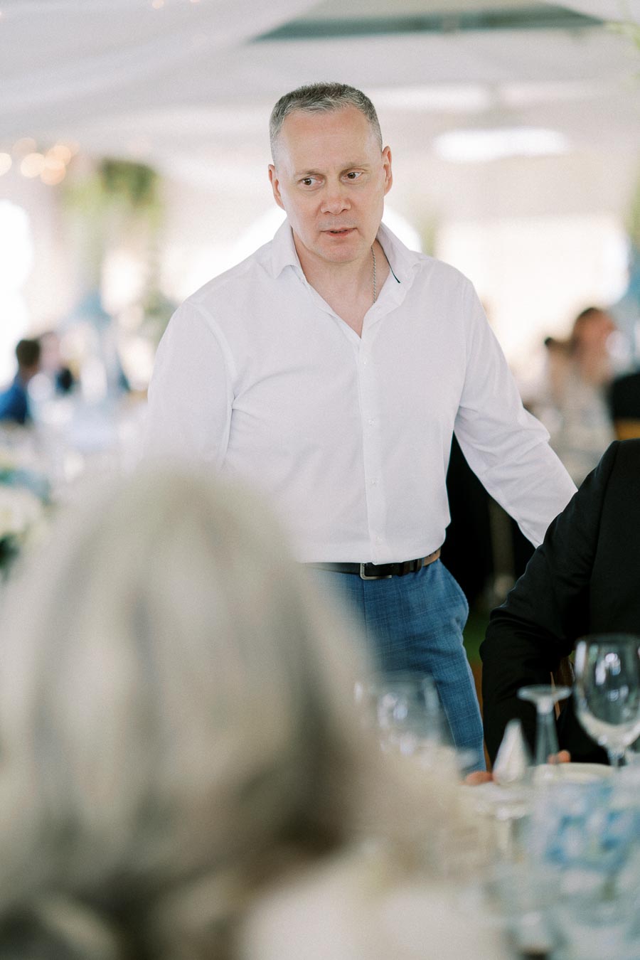 A man in a white shirt stands in a blurred indoor setting, engaged in conversation with blurred figures and table settings in the foreground, suggesting a social or formal event.