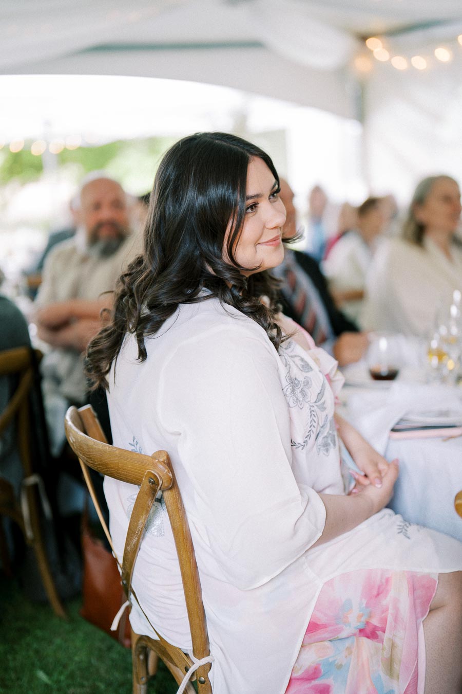 Smiling woman seated at an outdoor event, wearing a white floral dress, surrounded by other attendees under a canopy.