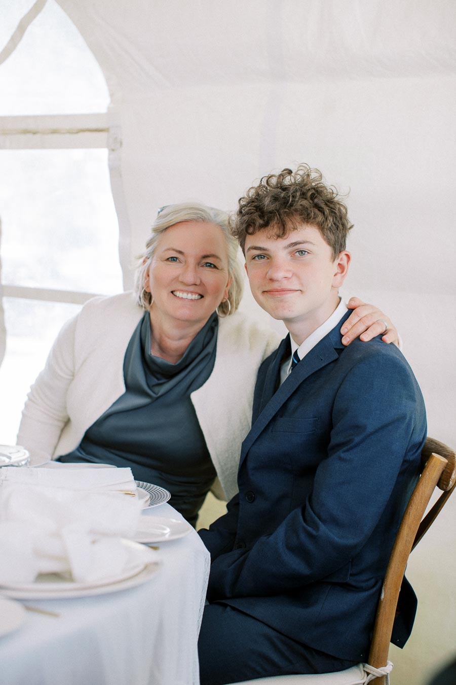 A smiling person with short white hair and a person with curly hair in a suit sitting together at a elegantly set dining table, inside a light-filled room.