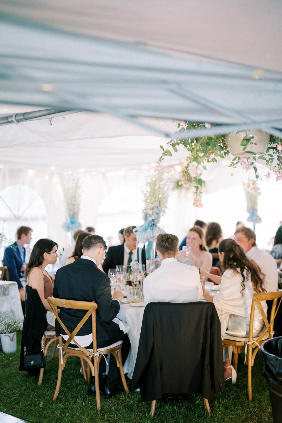 A group of elegantly dressed guests seated under a decorated tent at a wedding reception, enjoying a meal around a round table adorned with floral centerpieces.