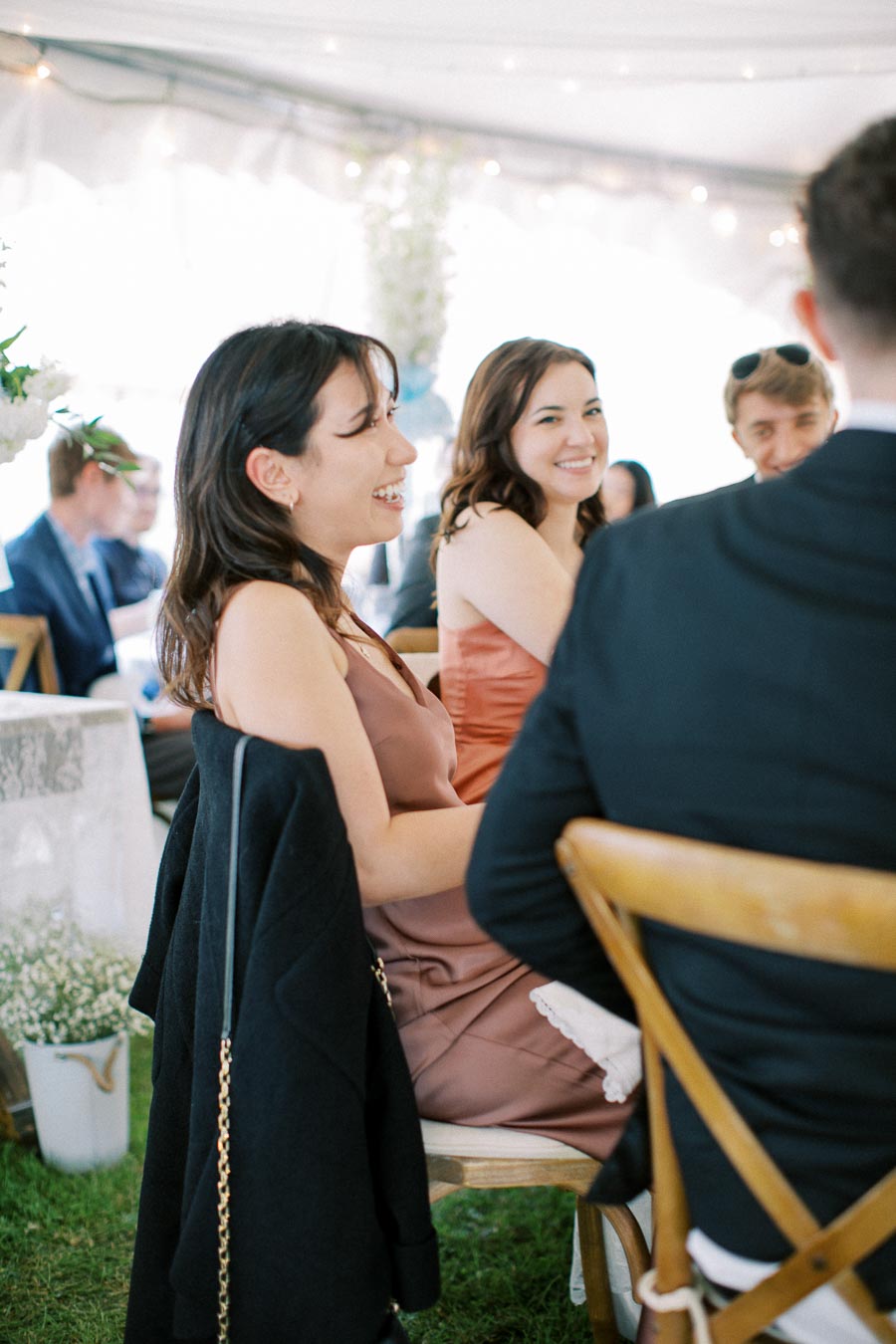 People smiling and socializing at a lively wedding reception, seated on wooden chairs with elegant floral decorations in the background.