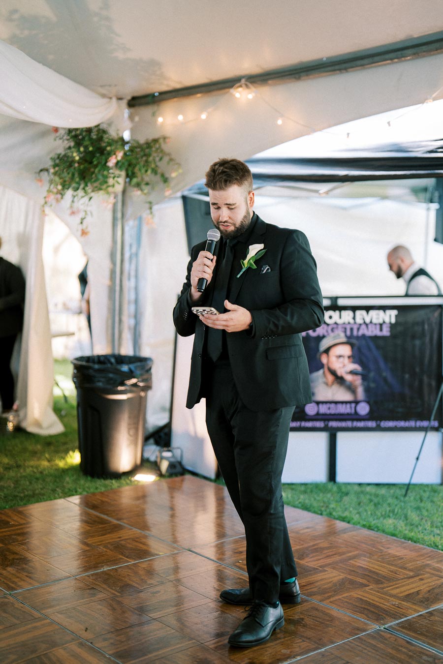 A man in a black suit holds a microphone while speaking at an outdoor event. The venue features decorative lighting and floral arrangements, and a poster in the background suggests entertainment is available for special occasions.