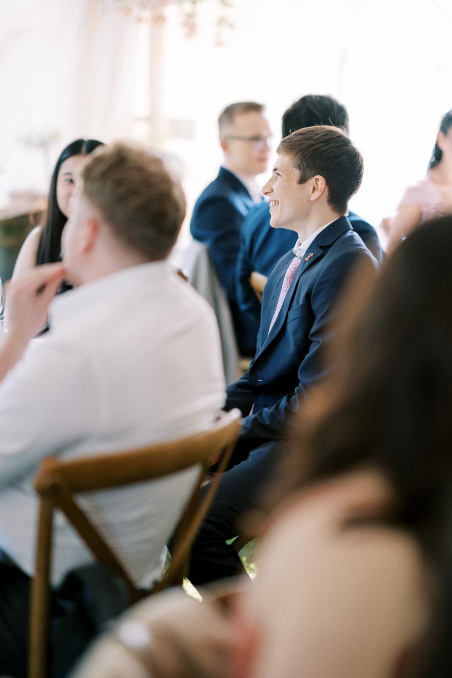 A group of people in formal attire seated at a wedding reception, with soft focus and natural lighting, capturing a joyous and elegant atmosphere.
