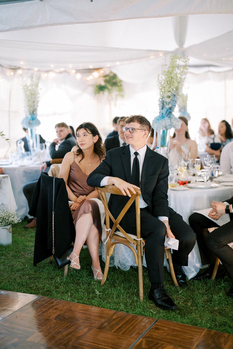 A couple sitting attentively at an elegant outdoor event, surrounded by elegantly decorated tables and floral arrangements under a white tent, suggesting a formal celebration or wedding reception.