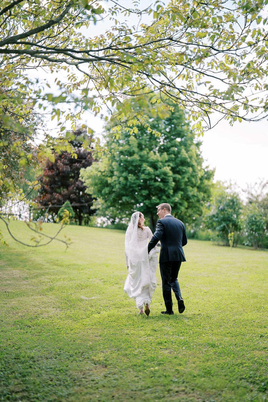 Newlywed couple walking hand in hand across a lush green lawn, surrounded by trees on a sunny day, capturing a romantic and serene outdoor wedding moment.
