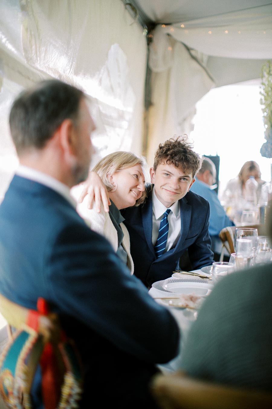 Young man in a suit smiling while sitting at a formal event, with a woman affectionately leaning on his shoulder.
